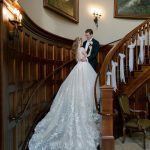Bride in lace ballgown and groom embracing on elegant curved wooden staircase with white ribbon decorations