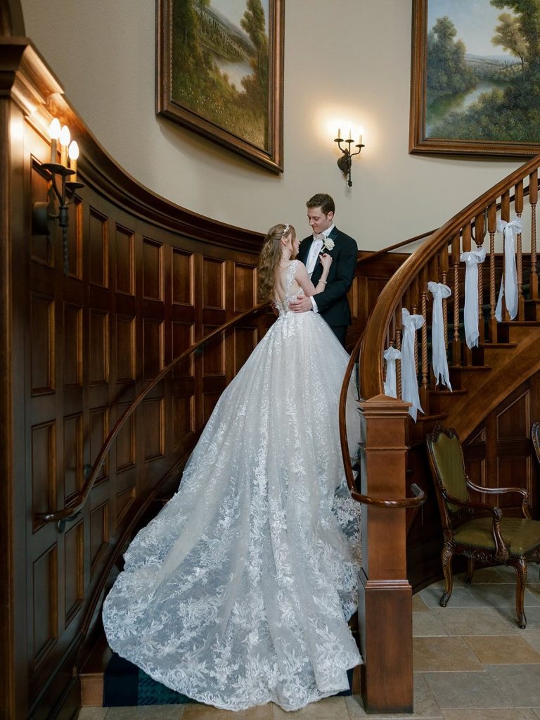 Bride in lace ballgown and groom embracing on elegant curved wooden staircase with white ribbon decorations