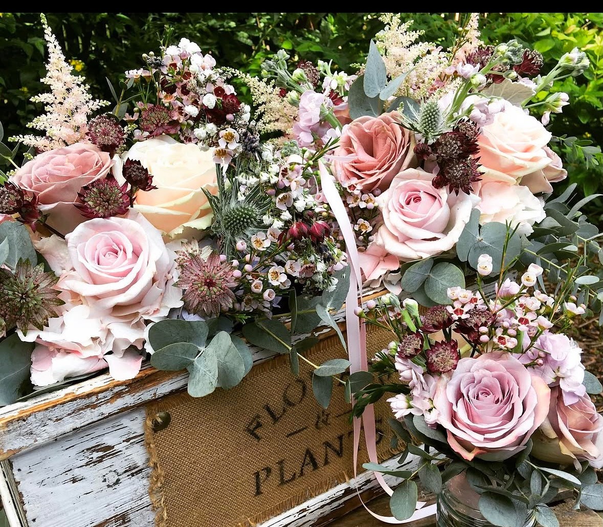 Wedding floral arrangement with blush roses, burgundy accents, and eucalyptus in rustic container with "thank you" tag