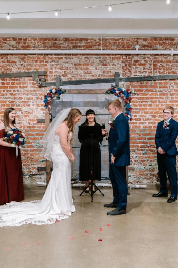 Wedding ceremony in industrial loft space with exposed brick wall, gray metal ceremony structure decorated with coral and blue flowers