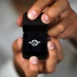 Hands holding open black velvet ring box displaying diamond engagement ring with center stone and side accents