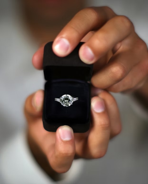 Hands holding open black velvet ring box displaying diamond engagement ring with center stone and side accents