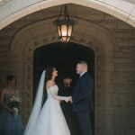 Bride and groom holding hands during ceremony under ornate stone archway with vintage lantern, bridesmaid visible to left
