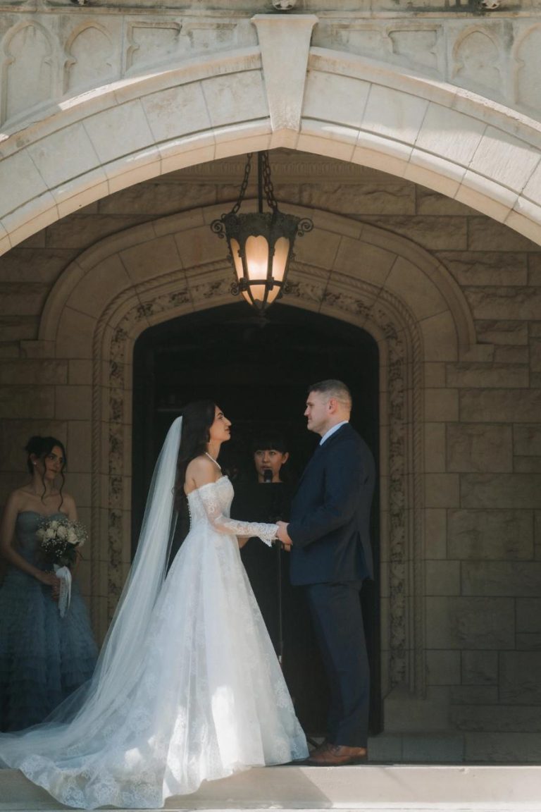 Bride and groom holding hands during ceremony under ornate stone archway with vintage lantern, bridesmaid visible to left