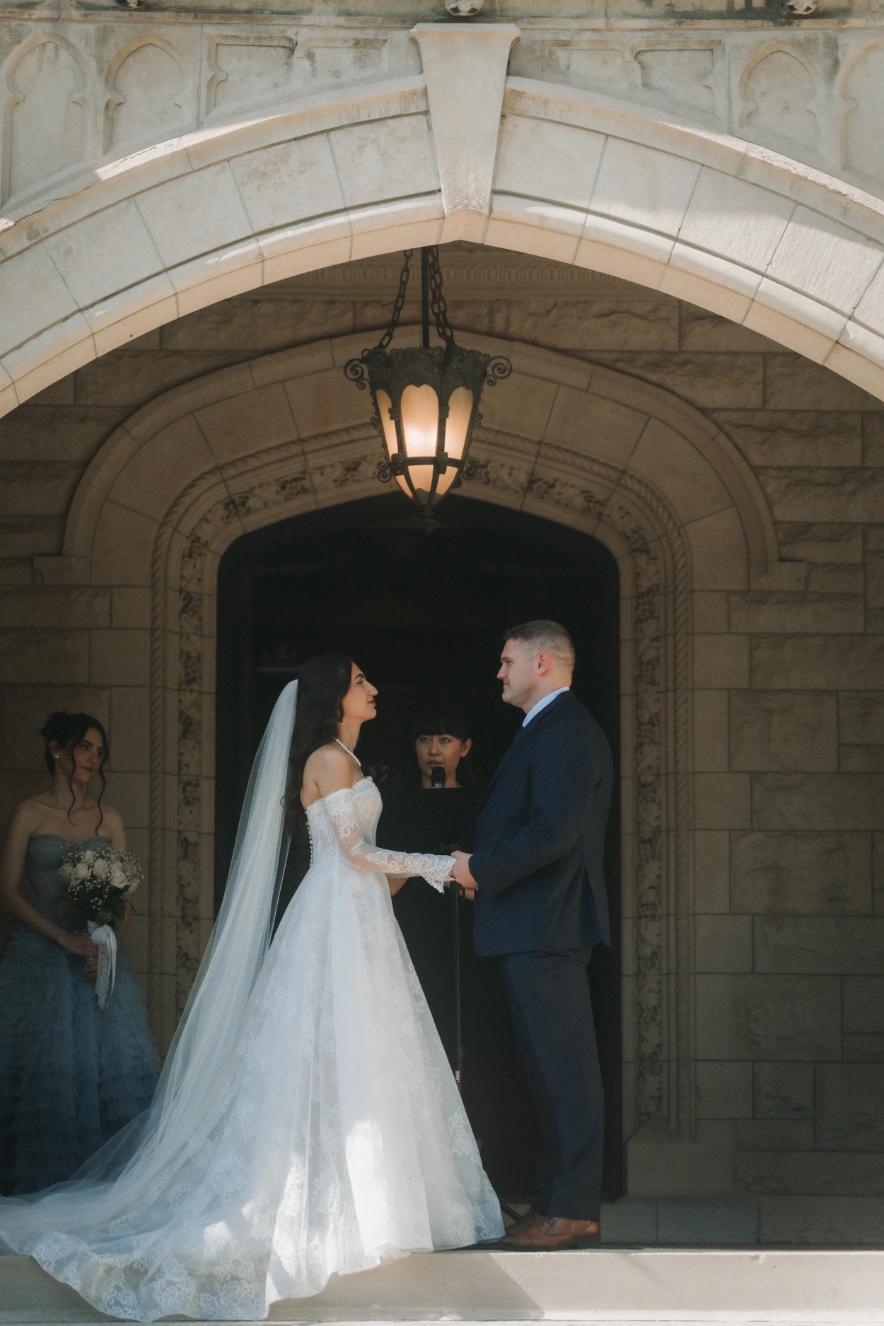 Bride and groom holding hands during ceremony under ornate stone archway with vintage lantern, bridesmaid visible to left