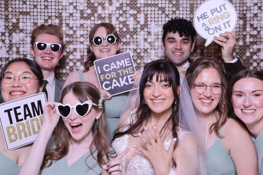 Wedding party posing with props in photo booth with sequin backdrop, bride showing engagement ring