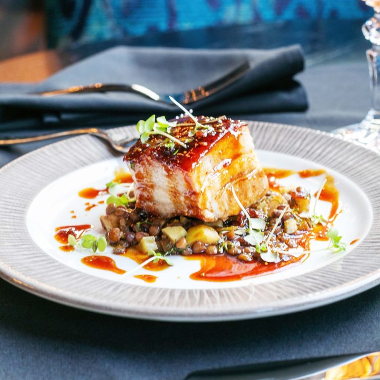 Glazed pork belly with crispy skin served over lentils, garnished with microgreens and sauce drizzle