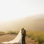 Bride and groom embracing in misty mountain landscape at golden hour with flowing cathedral veil