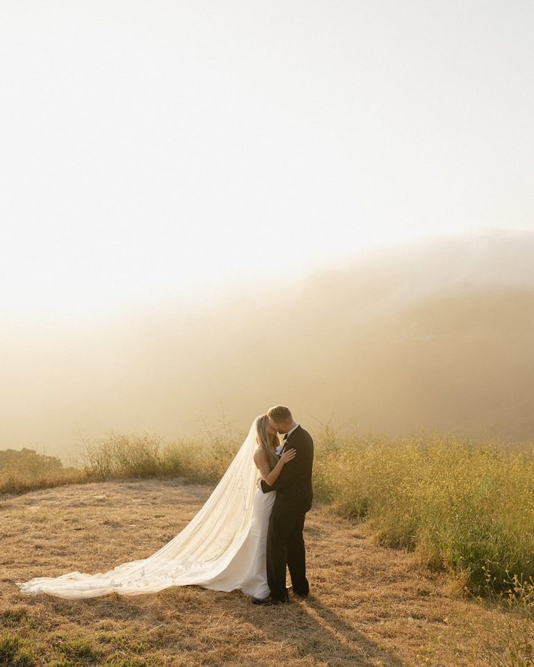 Bride and groom embracing in misty mountain landscape at golden hour with flowing cathedral veil