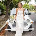 Bride in white gown holding soft pink and white bouquet, leaning against vintage white car