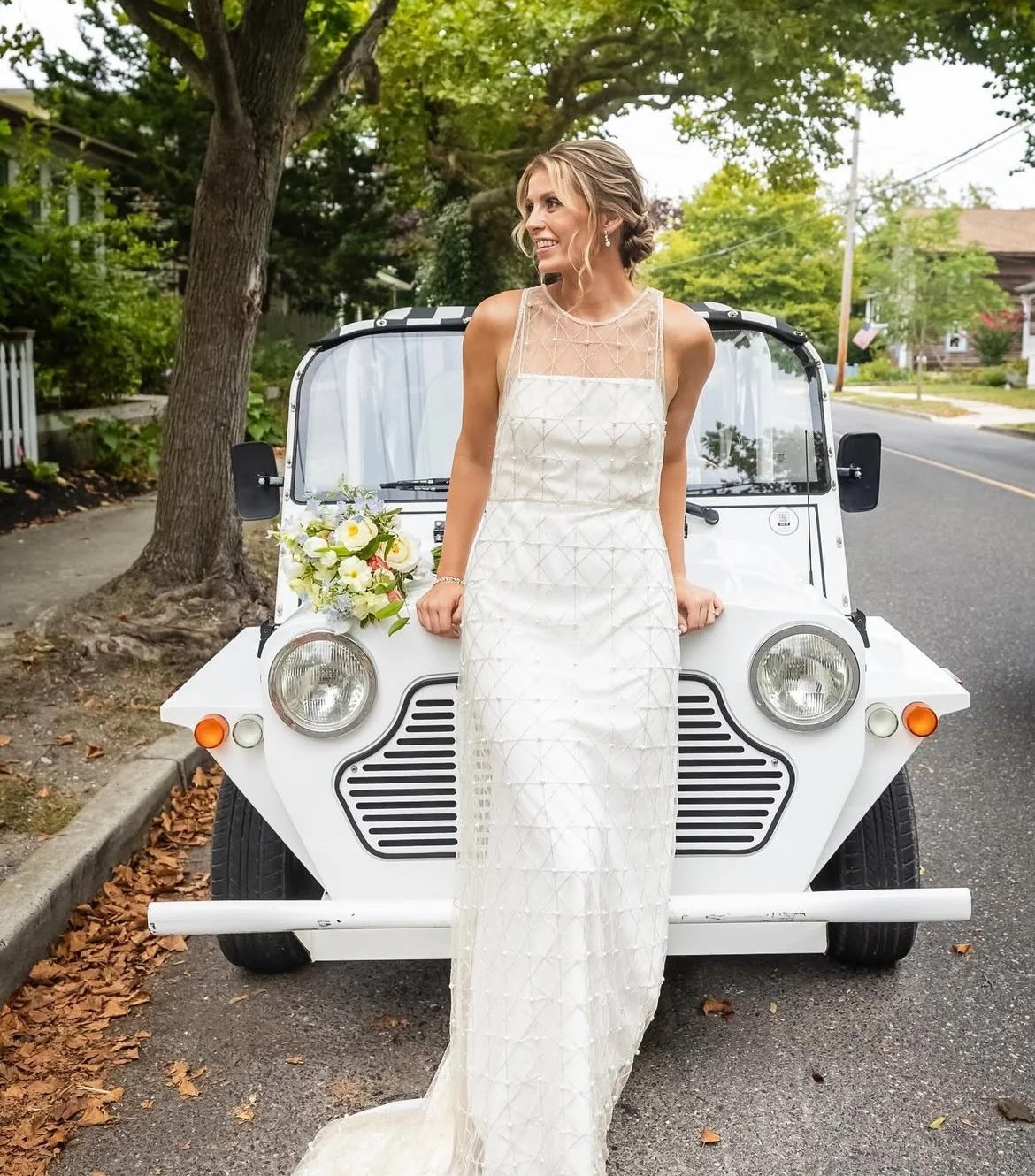 Bride in white gown holding soft pink and white bouquet, leaning against vintage white car