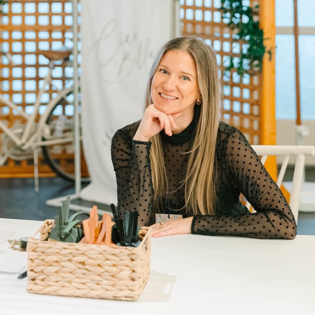 Smiling woman in polka dot black blouse seated at white desk with wicker basket of wedding supplies in bright studio space