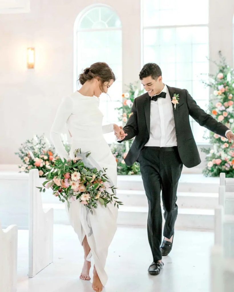 Newlyweds walking down aisle hand in hand in bright white ceremony space with pink floral arrangements