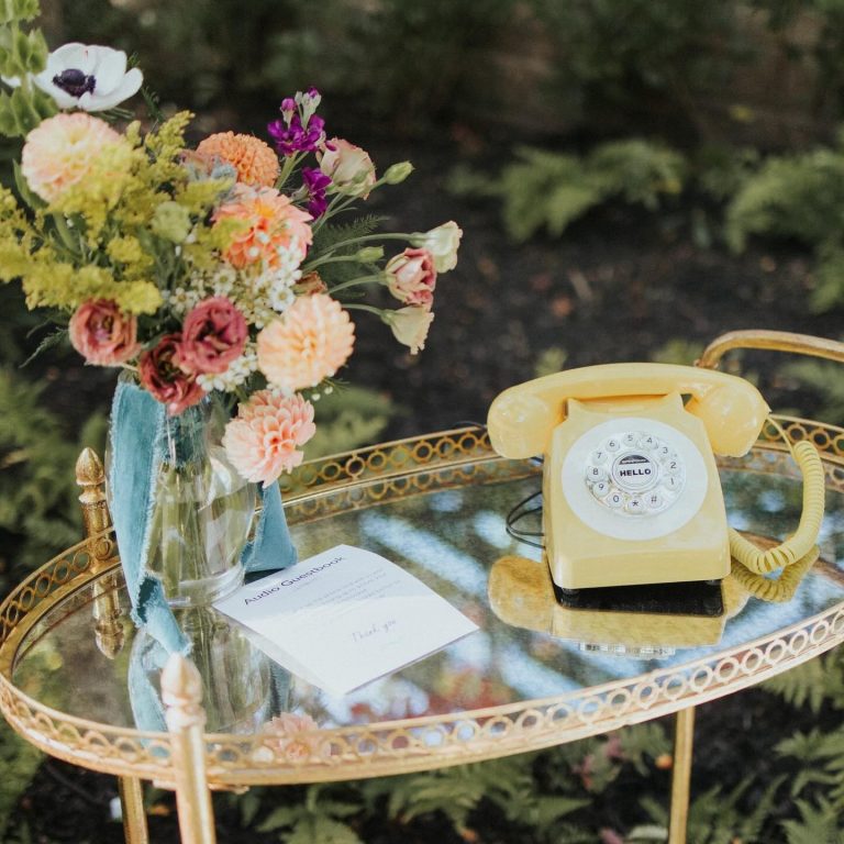Vintage gold mirrored tray displaying colorful dahlia and anemone bouquet beside yellow rotary phone and wedding guestbook