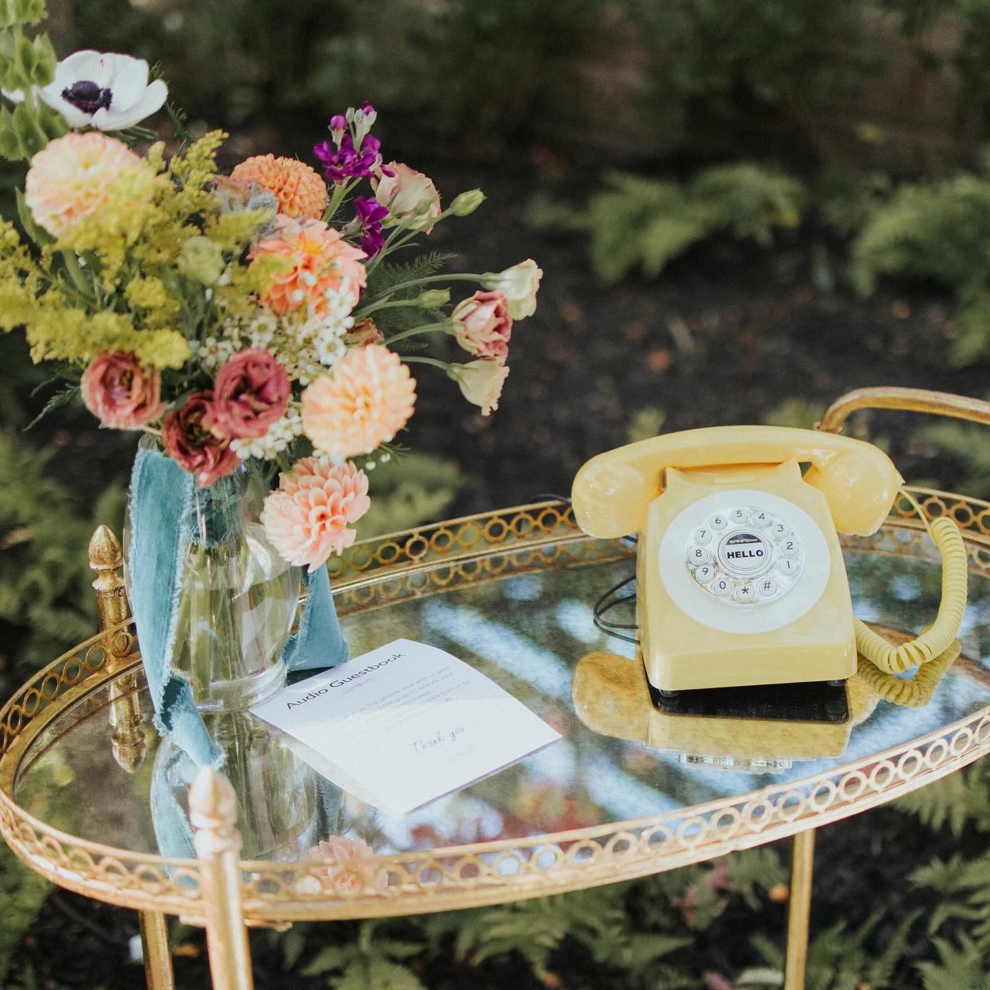 Vintage gold mirrored tray displaying colorful dahlia and anemone bouquet beside yellow rotary phone and wedding guestbook