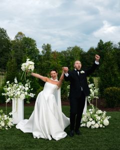 Newlyweds cheering with clasped hands and raised fists at outdoor ceremony with white florals