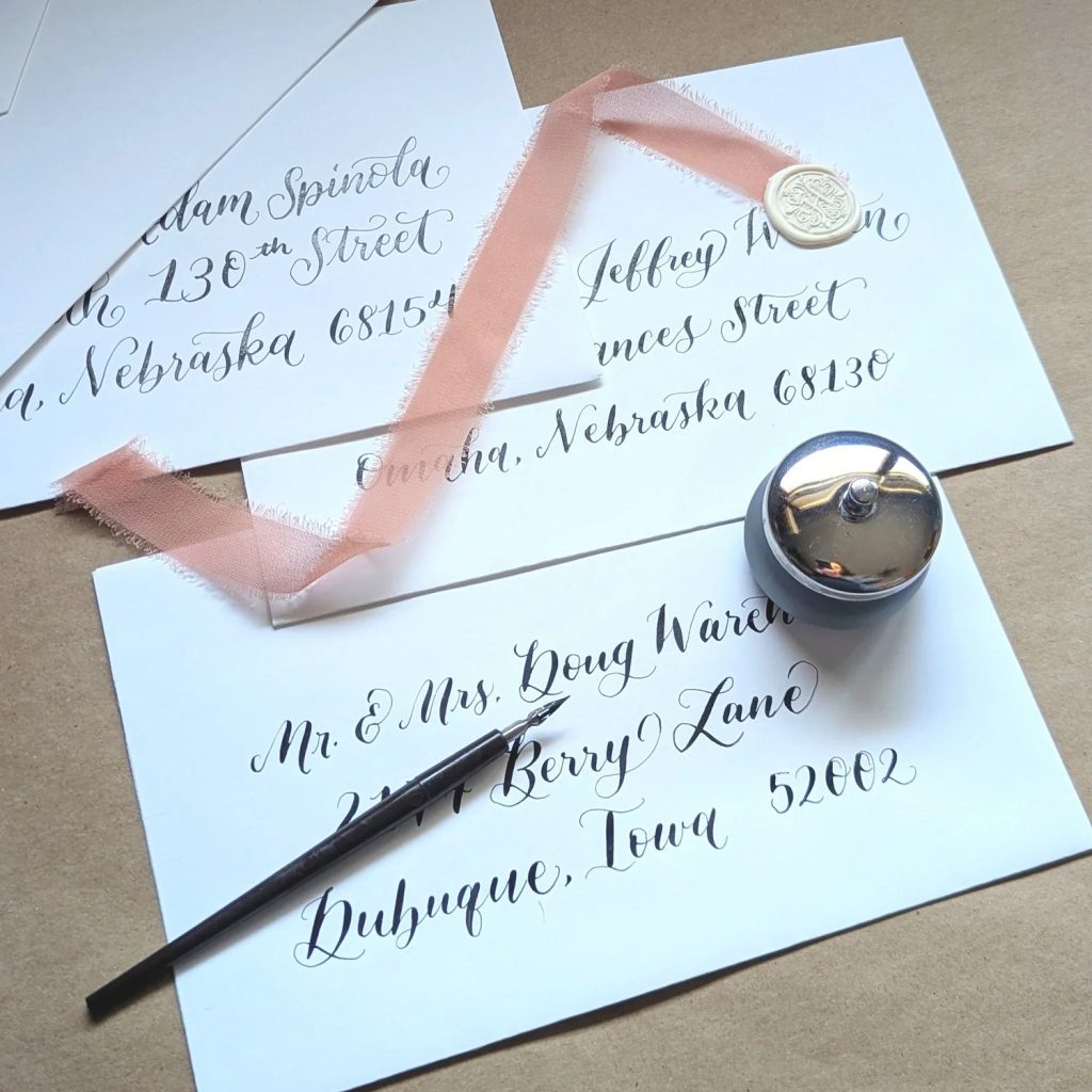 Hand-addressed wedding envelopes with elegant calligraphy featuring Nebraska addresses, displayed with pink silk ribbon and calligraphy pen