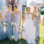Bride in white gown with bridesmaids in blue dresses holding white and blush bouquets at outdoor wedding venue