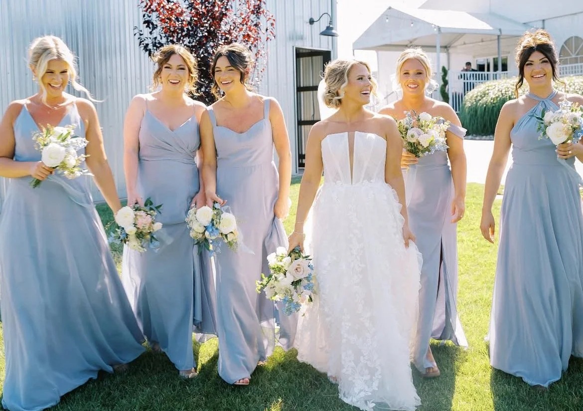 Bride in white gown with bridesmaids in blue dresses holding white and blush bouquets at outdoor wedding venue