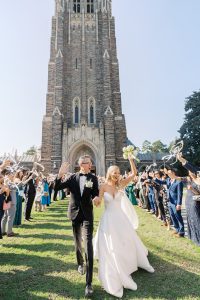 Bride and groom exit historic stone chapel tower through enthusiastic ribbon wand send-off line