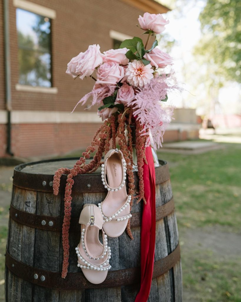 Romantic wedding bouquet with blush roses, trailing amaranthus, and bridal heels displayed on weathered barrel outdoors