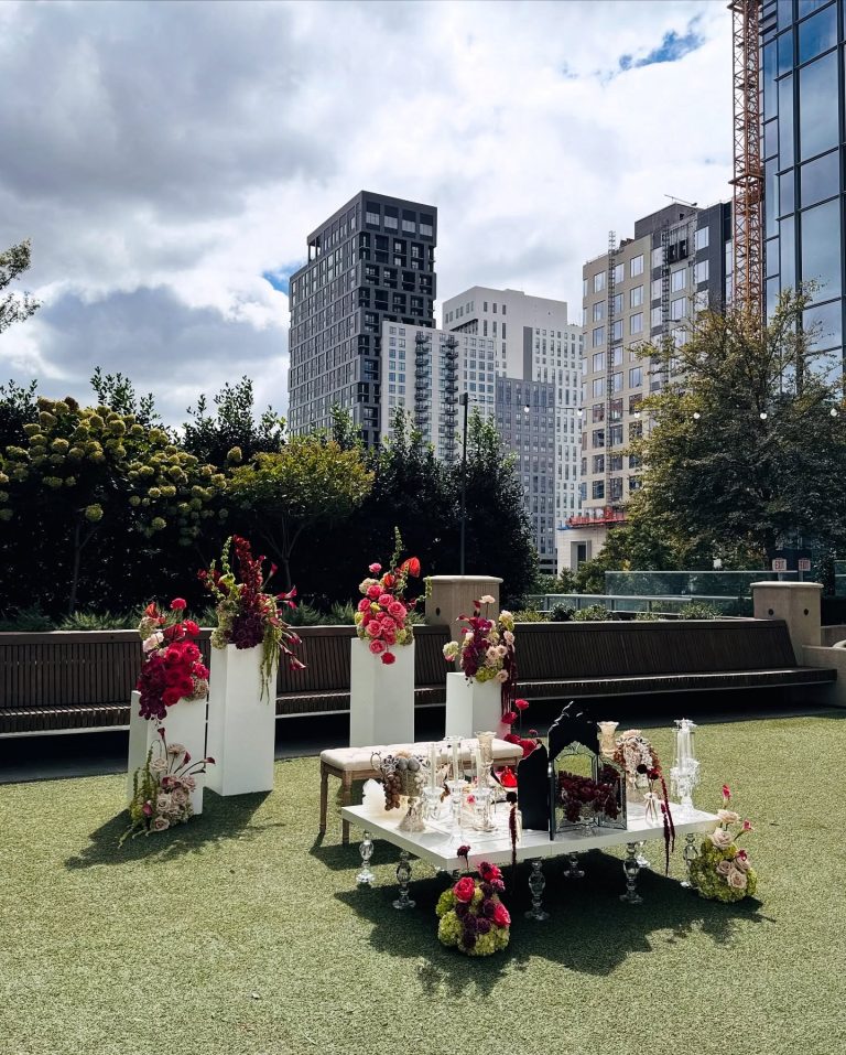 Persian Sofreh Aghd wedding spread with white ceremonial table, pink and burgundy floral arrangements on pillars, and urban rooftop backdrop