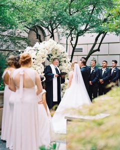 Outdoor wedding ceremony with white floral arch, officiant, couple, and wedding party under green trees