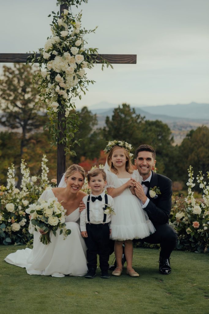 Bride and groom with two children posing beneath a wooden ceremony arch adorned with white roses, hydrangeas, and greenery, with mountain views in the background.