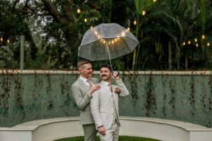 Two grooms in cream suits sharing umbrella under string lights with lush tropical greenery backdrop