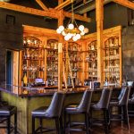 Rustic bar interior with exposed wooden beams, globe chandelier, and leather bar stools facing illuminated liquor shelves
