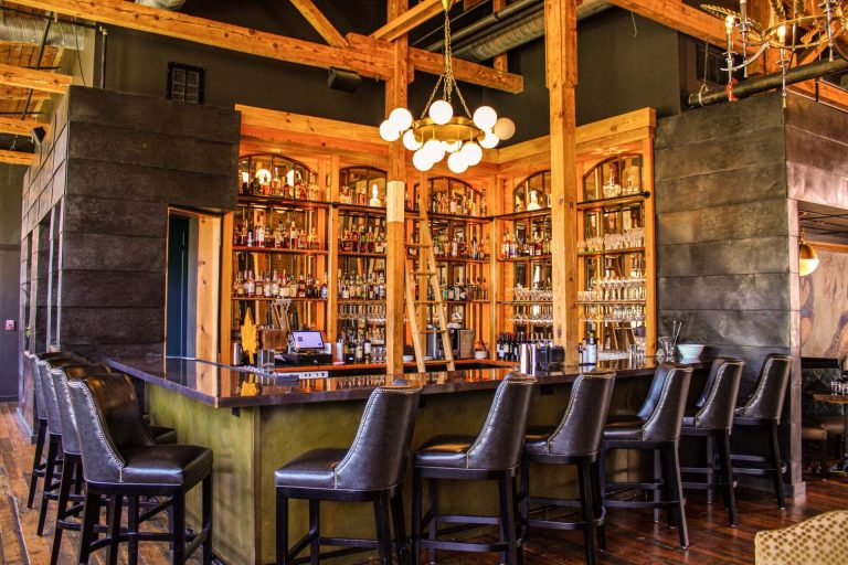 Rustic bar interior with exposed wooden beams, globe chandelier, and leather bar stools facing illuminated liquor shelves
