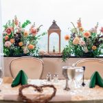 Rustic wedding reception tablescape with coral and peach floral centerpieces flanking copper lantern