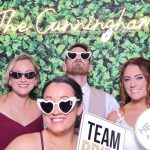 Wedding guests posing with novelty sunglasses and "Team Groom" and "He Put a Ring on It" signs against greenery wall backdrop