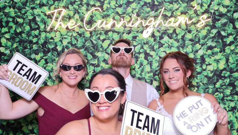 Wedding guests posing with novelty sunglasses and "Team Groom" and "He Put a Ring on It" signs against greenery wall backdrop