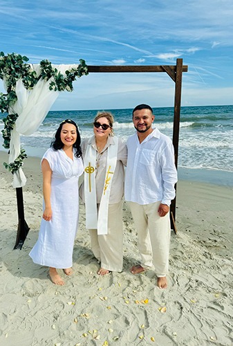 Wedding officiant with couple at beachfront ceremony under natural arbor