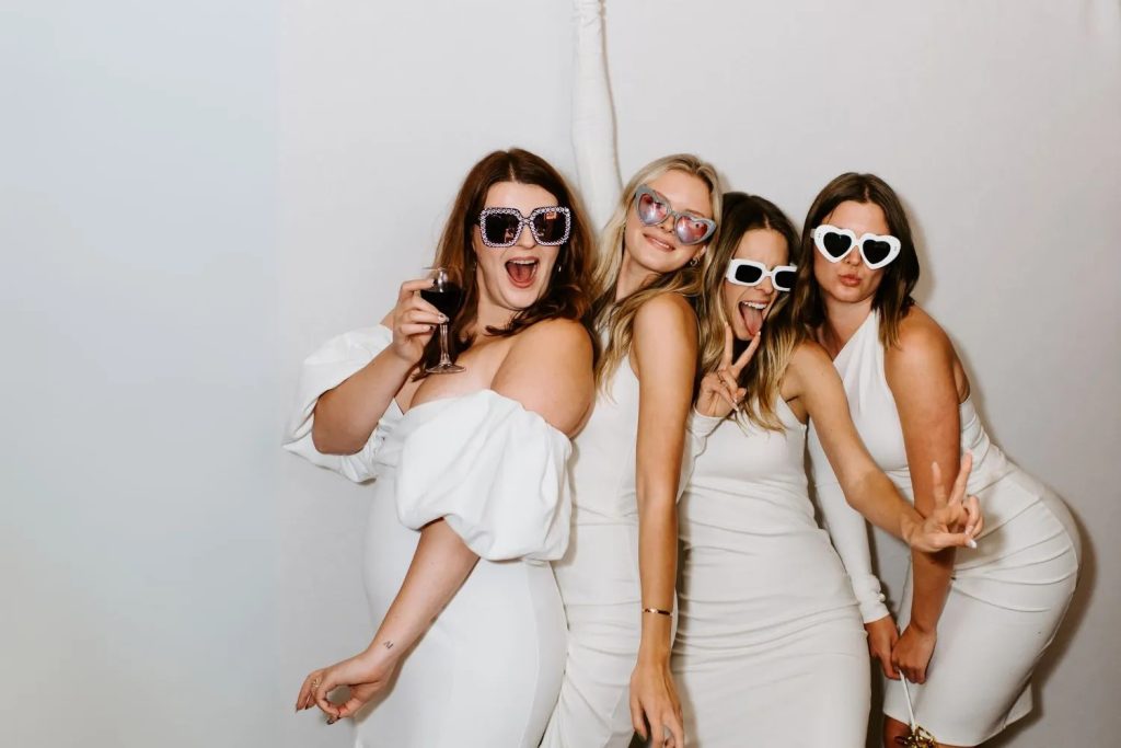 Four women in white dresses wearing playful sunglasses in photo booth
