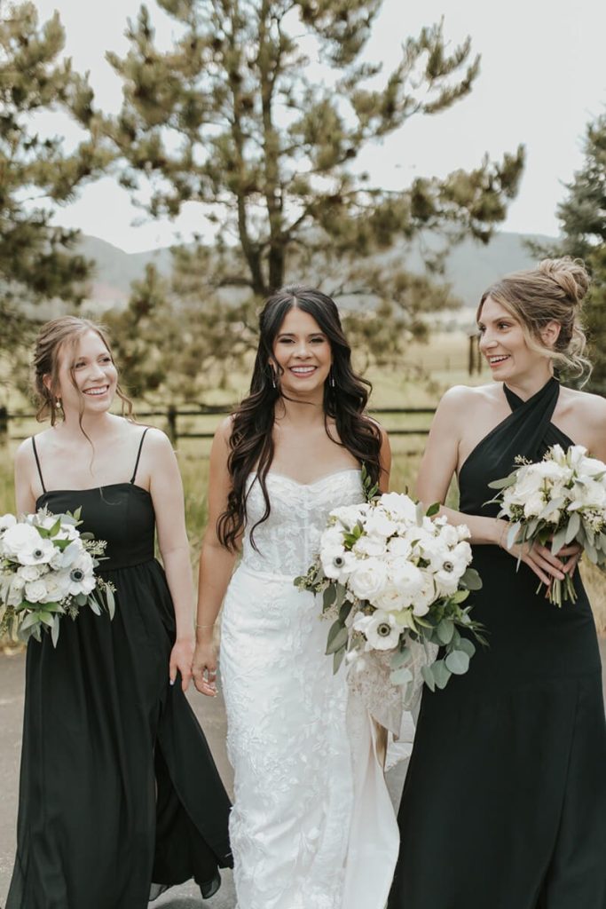 Bride in white lace gown with two bridesmaids in black dresses holding white bouquets at outdoor Denver wedding