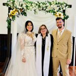 Bride and groom with Reverend Susie beneath chandelier and sunflower-accented floral arch
