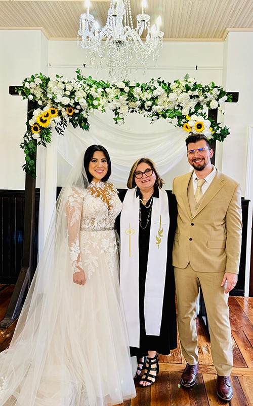 Bride and groom with Reverend Susie beneath chandelier and sunflower-accented floral arch