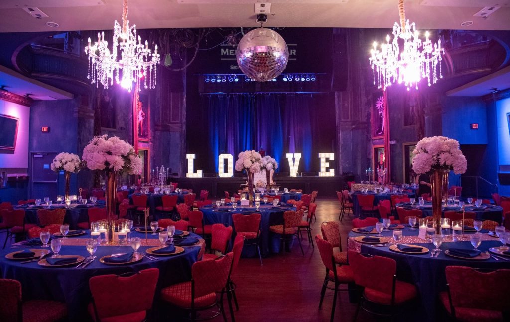 Dramatic ballroom reception with illuminated LOVE sign, crystal chandeliers, red chairs, and tall white floral centerpieces