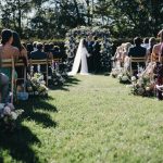 Wedding ceremony aisle lined with lavender and white floral arrangements on wooden chairs leading to couple at floral arch