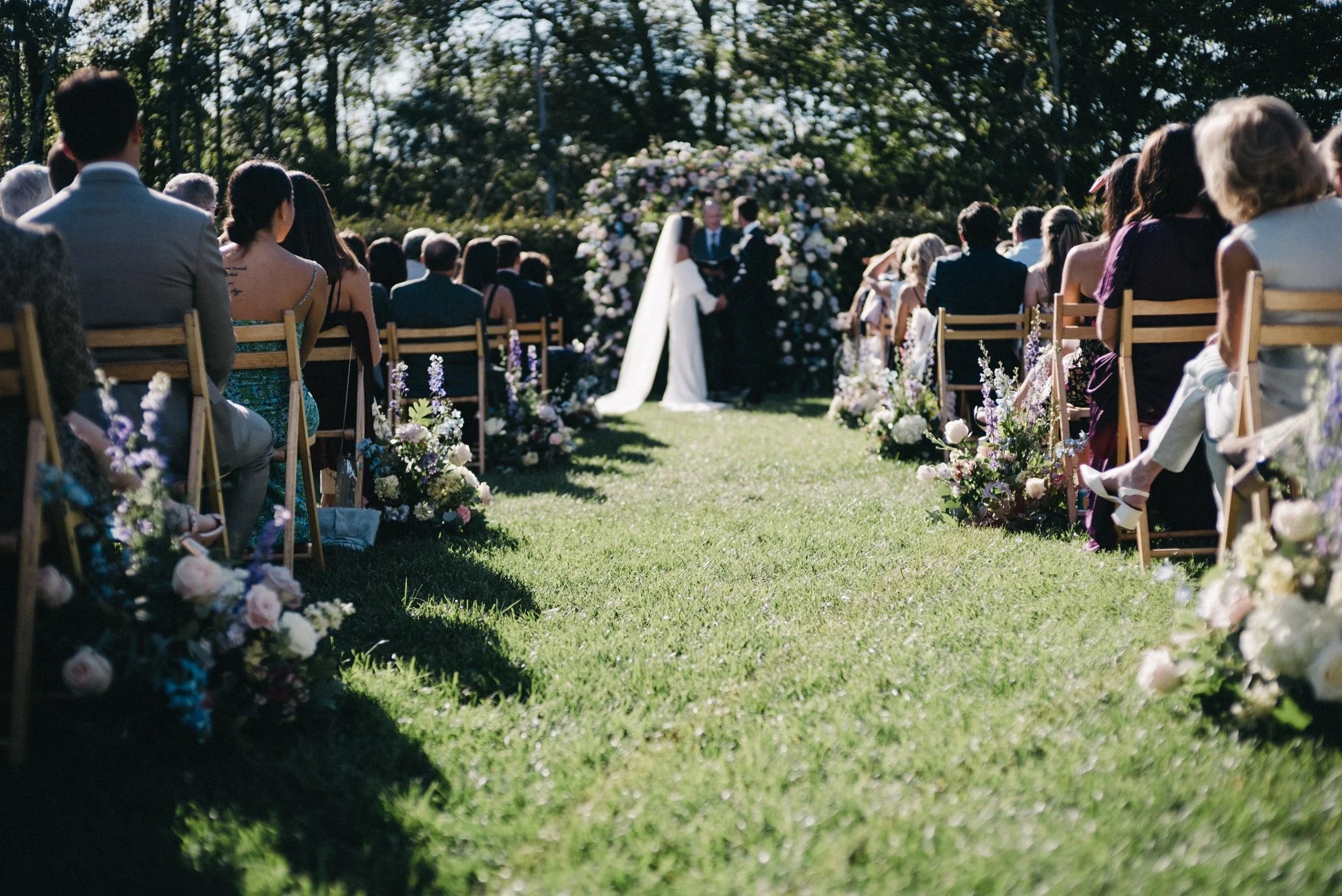 Wedding ceremony aisle lined with lavender and white floral arrangements on wooden chairs leading to couple at floral arch