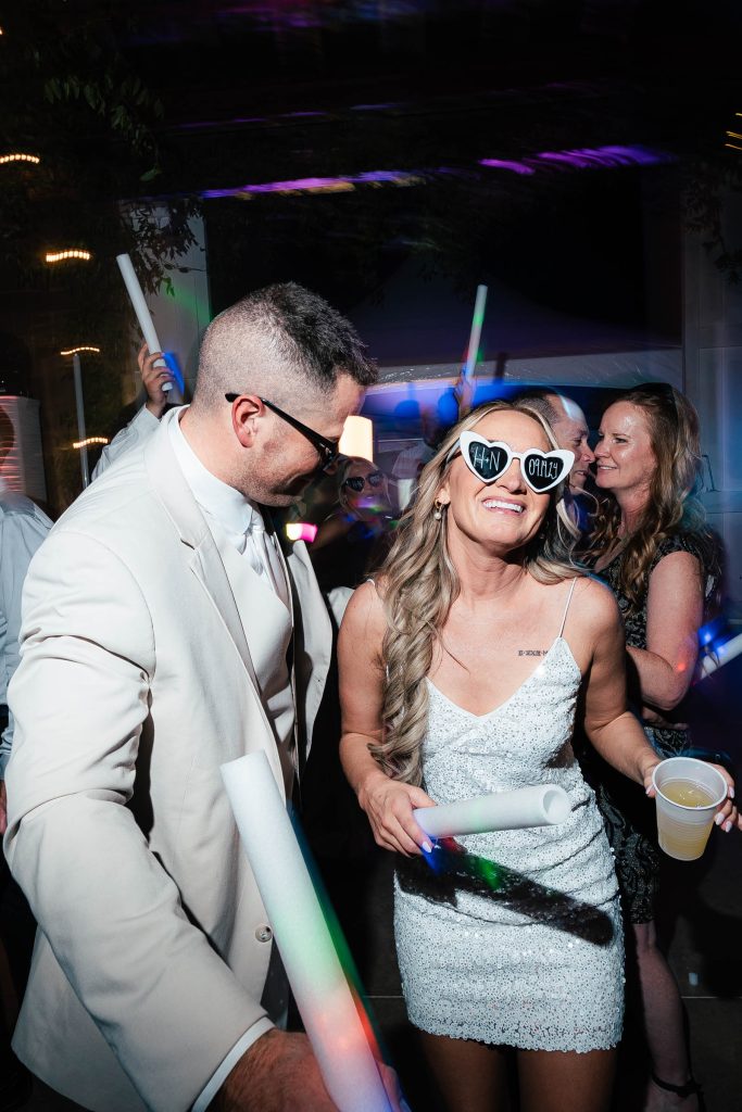 Bride and groom celebrating at wedding reception with glow sticks and "TEAM BRIDE" sunglasses under purple lighting