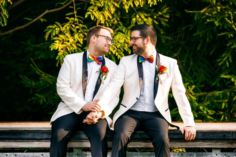 Two grooms in white tuxedo jackets with rainbow bow ties and orange boutonnieres sit together on a wooden bench against vibrant green foliage