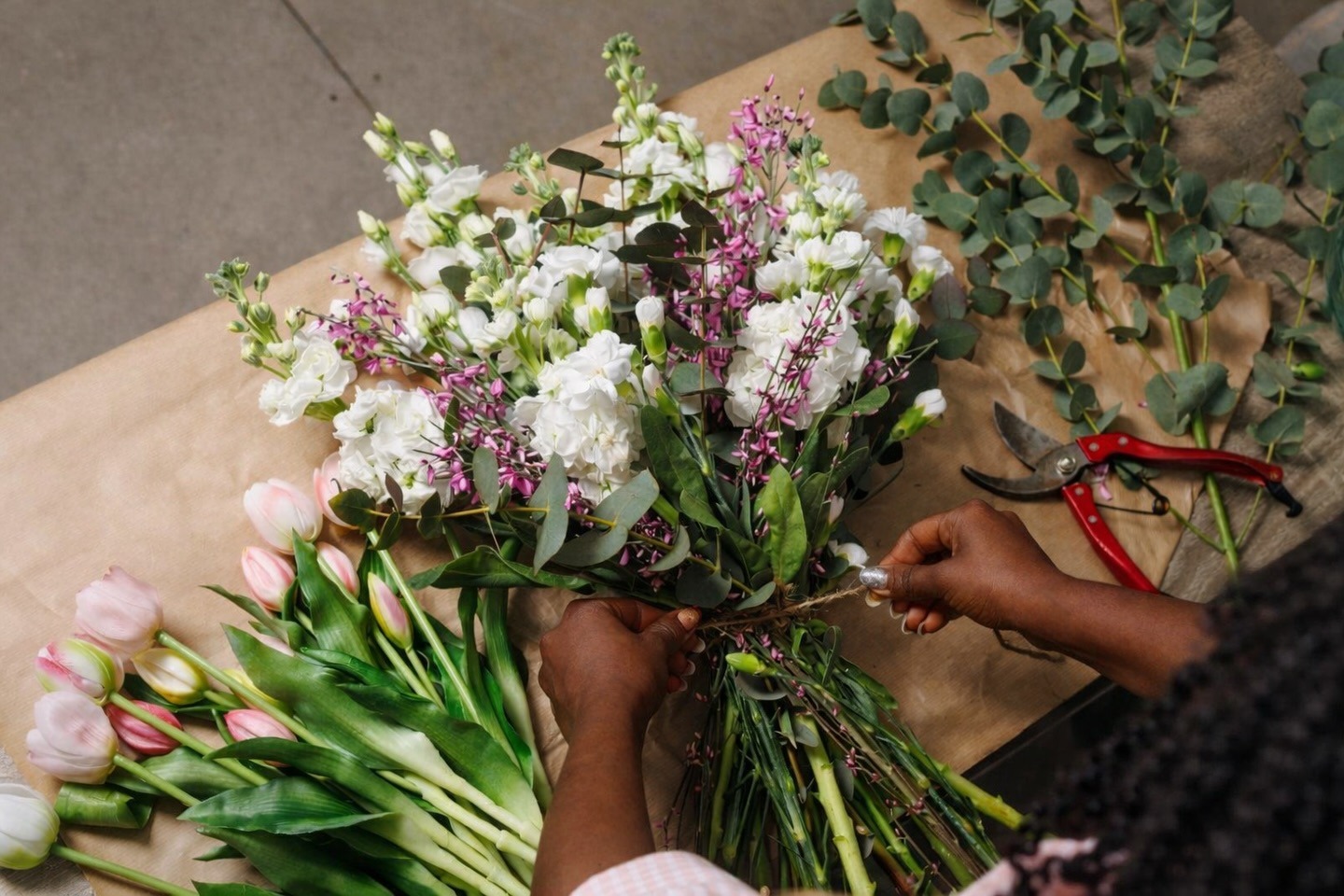 Florist binding stems of white and purple wedding flowers with pink tulips and eucalyptus beside red pruning shears