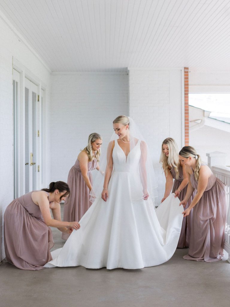 Bridesmaids in mauve dresses adjusting bride Catherine's voluminous wedding gown train on covered porch