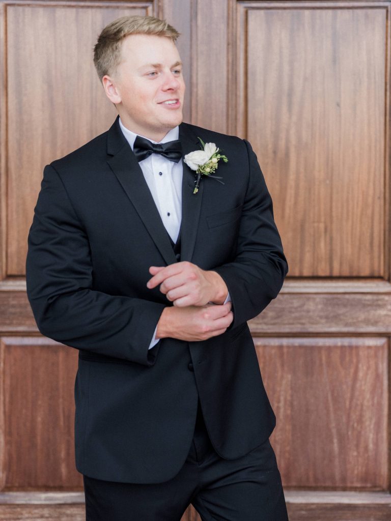Groom in black tuxedo with bow tie and white boutonniere against wood-paneled wall
