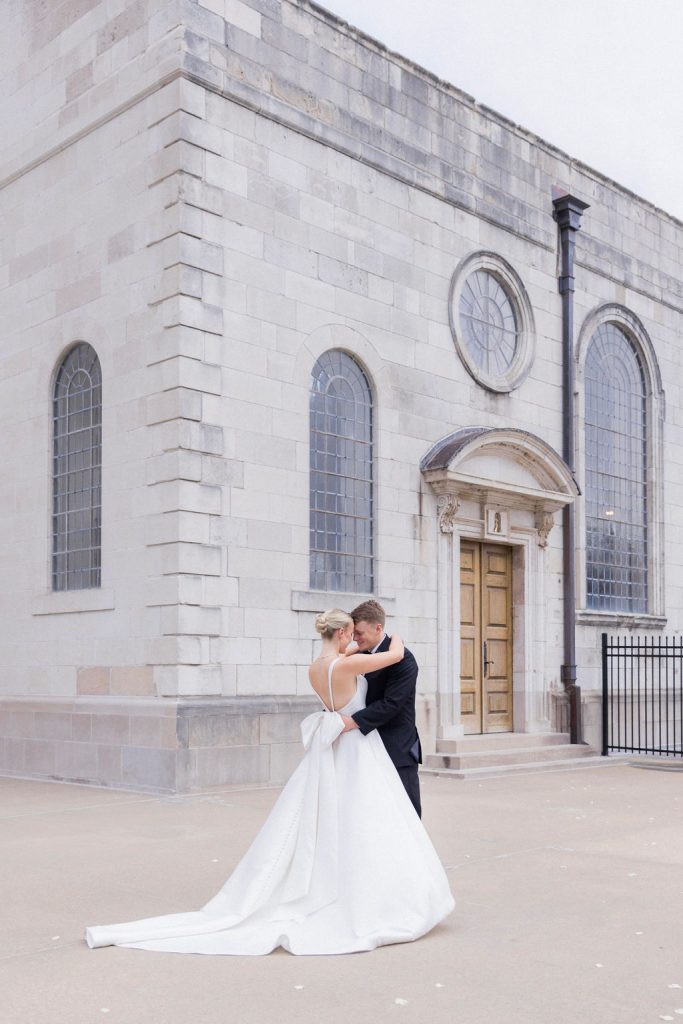 Catherine and Zach embrace outside a historic stone church with arched windows, the bride's gown flowing behind them