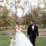 Newlywed couple holding hands while walking across manicured lawn with autumn trees