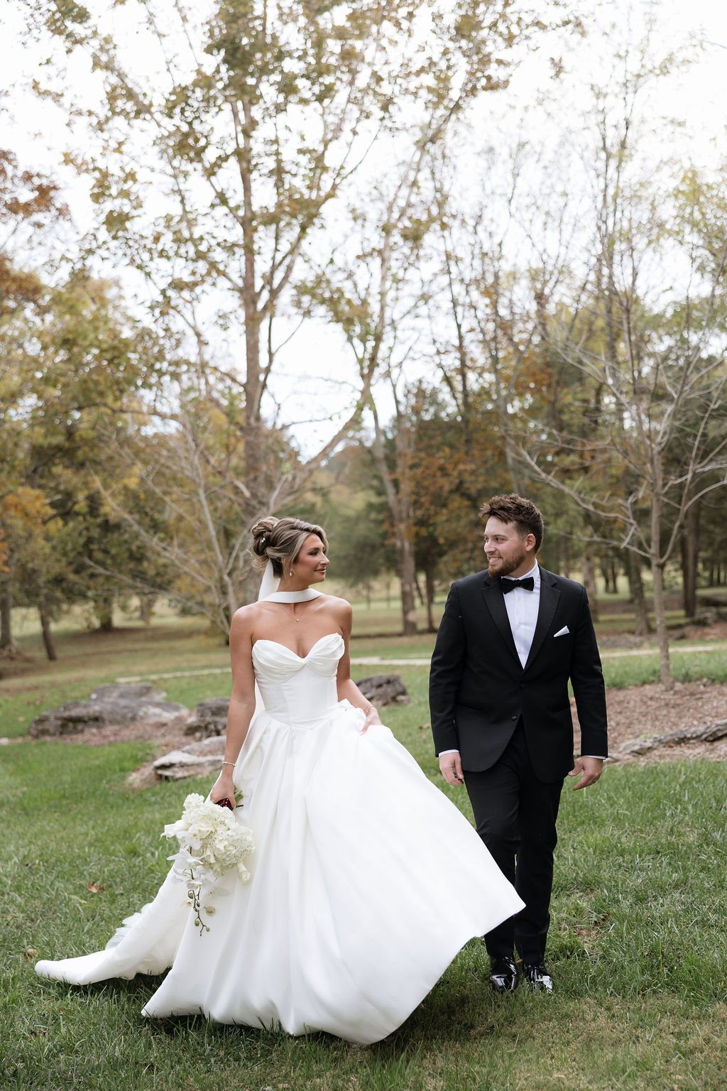 Newlywed couple holding hands while walking across manicured lawn with autumn trees